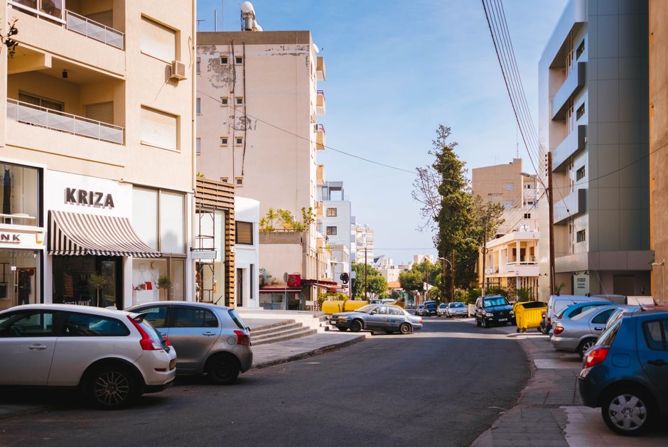 A modern public bus driving through a sunny street in Cyprus, with palm trees and buildings in the background.